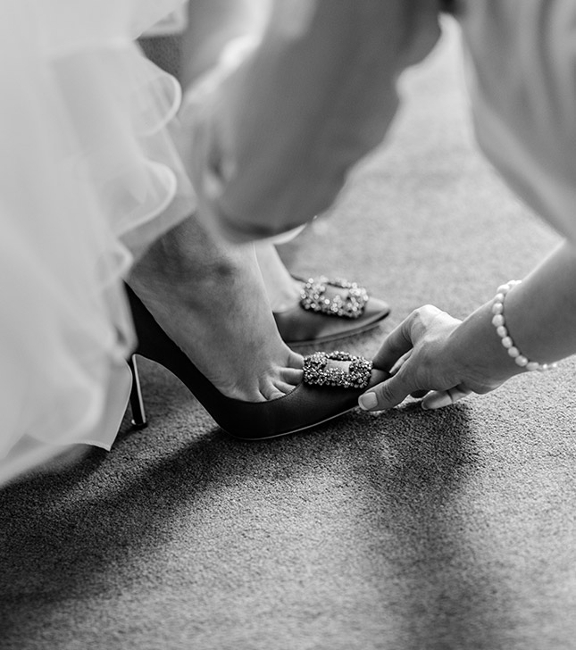In a black and white image captured by a Surrey wedding photographer, an elegant, embellished high-heeled shoe is gently adjusted by another with a bracelet on their wrist. The carpeted floor and part of a flowing dress complete this documentary-style moment.