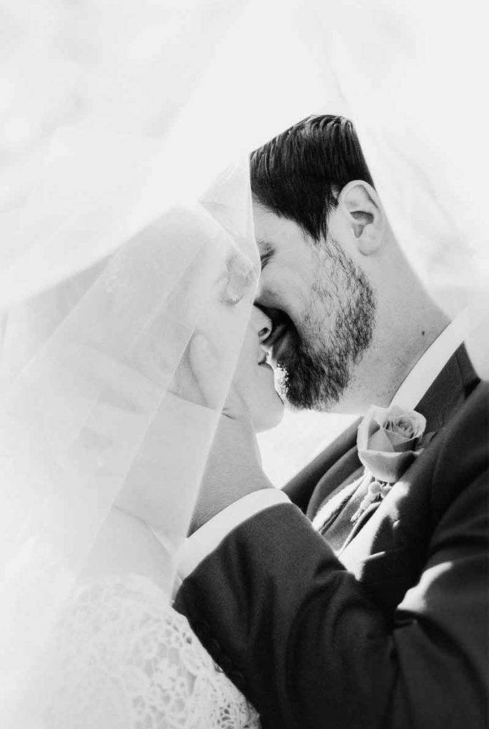 A black and white image captured by a Surrey wedding photographer shows a bride and groom under a veil, sharing an intimate moment. The groom has a beard while the bride's dress features intricate lace. Both have their eyes closed, poised for an impending kiss.
