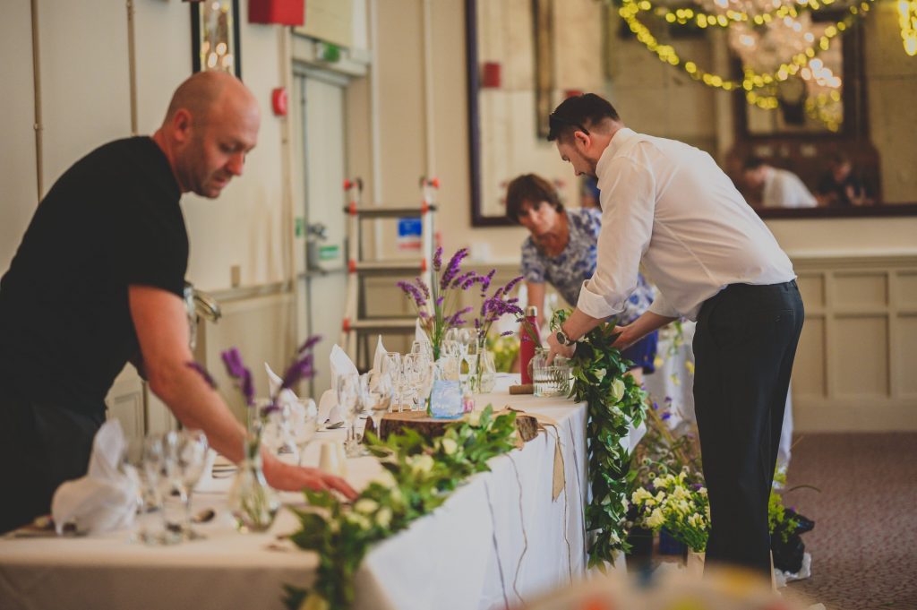 Two people are setting up a decorated table in a well-lit room. One arranges floral garlands, while the other adjusts glassware and napkins. Observed by a third person in the background, the elegant decor includes flowers and string lights—a perfect moment for any documentary wedding photographer in Surrey to capture.