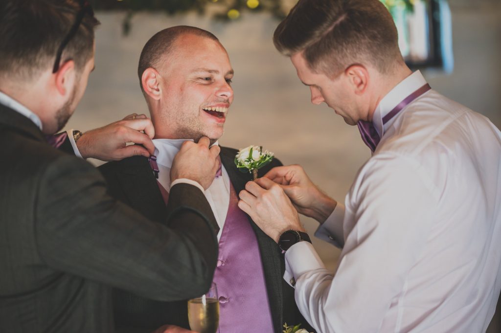 Three men are in a room, dressed in formal attire. The man in the center is smiling as the other two adjust his tie and boutonniere. He holds a glass of champagne. A documentary wedding photographer from Surrey captures the festive scene, likely preparing for a wedding or formal event.