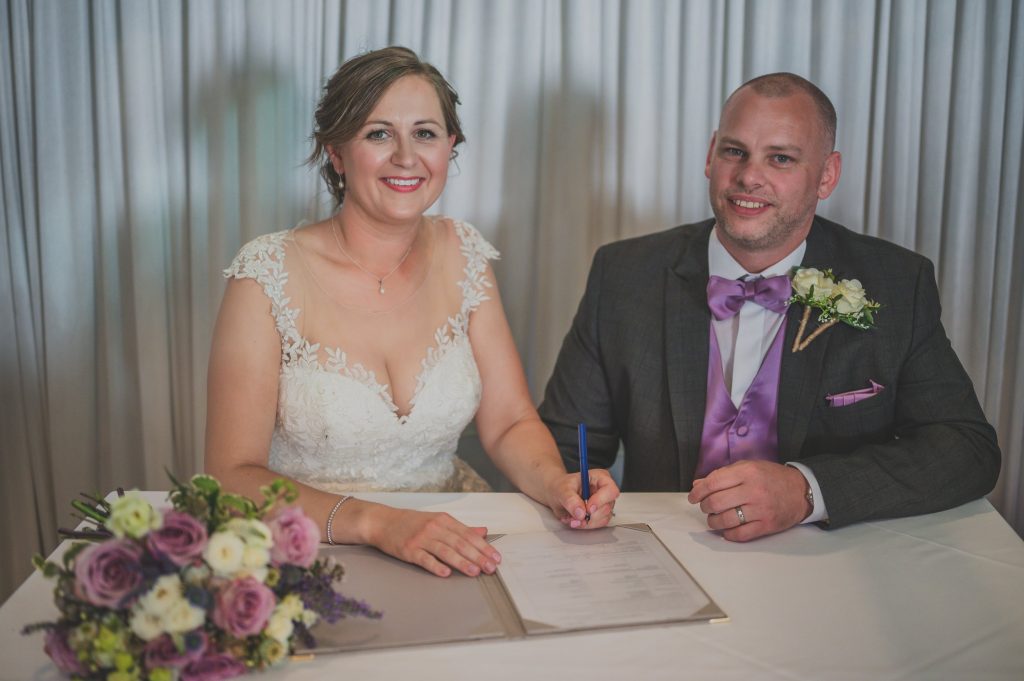 A smiling bride in a lace wedding dress and groom in a gray suit with a purple tie sit at a table while the bride signs a document. A bouquet of flowers lies on the table in front of them. Captured beautifully by a documentary wedding photographer Surrey, curtains serve as the backdrop.