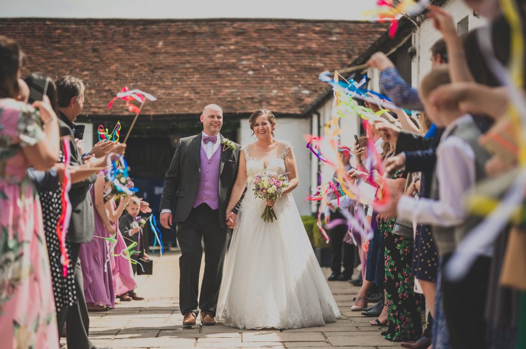 A bride and groom walk hand-in-hand down an aisle lined with joyful guests, captured perfectly by a documentary wedding photographer in Surrey. The bride in her white dress holds a bouquet, while the groom sports a vest with a bow tie. Guests wave colorful streamers, creating a festive outdoor atmosphere.