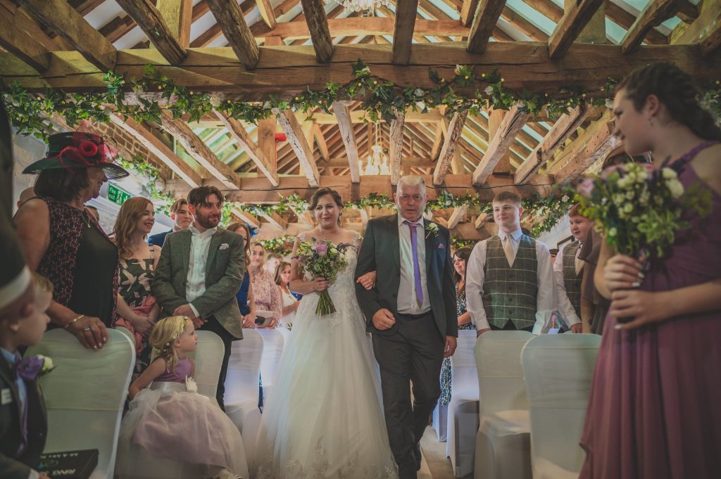 The bride, holding a bouquet, walks down the aisle with an older man, possibly her father, in a rustic, ivy-draped venue. Guests watch on both sides, including children, and a bridesmaid in a purple dress holds flowers. The groom stands to the side, smiling as the wedding photographer from Surrey captures every precious moment.