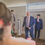 A man in a suit, smiling and holding sunglasses, enters a room with two other men in suits. A person with flower-adorned hair is in the foreground, blurred, adding an artistic touch reminiscent of a documentary wedding photographer's style. The room has white walls and a wooden floor.