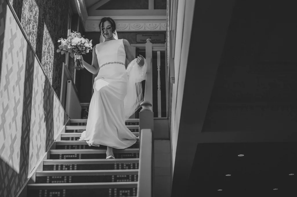 A bride in a long gown descends a staircase gracefully, holding a bouquet of flowers. Sunlight casts patterned shadows on the wall, creating a dramatic atmosphere expertly captured by a wedding photographer in Surrey. The black and white effect adds a timeless quality to the scene.