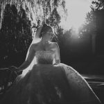 Black and white photo of a bride in a strapless gown with floral patterns, leaning against a car. As captured by a documentary wedding photographer in Surrey, she wears a veil and sunglasses, looking to the side. Sunlight creates dramatic contrasts with shadows from surrounding trees.