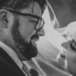 A black and white close-up of a bride and groom smiling at each other, captured by a Surrey wedding photographer. The groom, wearing glasses and a beard, gazes adoringly at the bride, whose face is partially covered by a veil, adding an enchanting touch to this romantic moment.