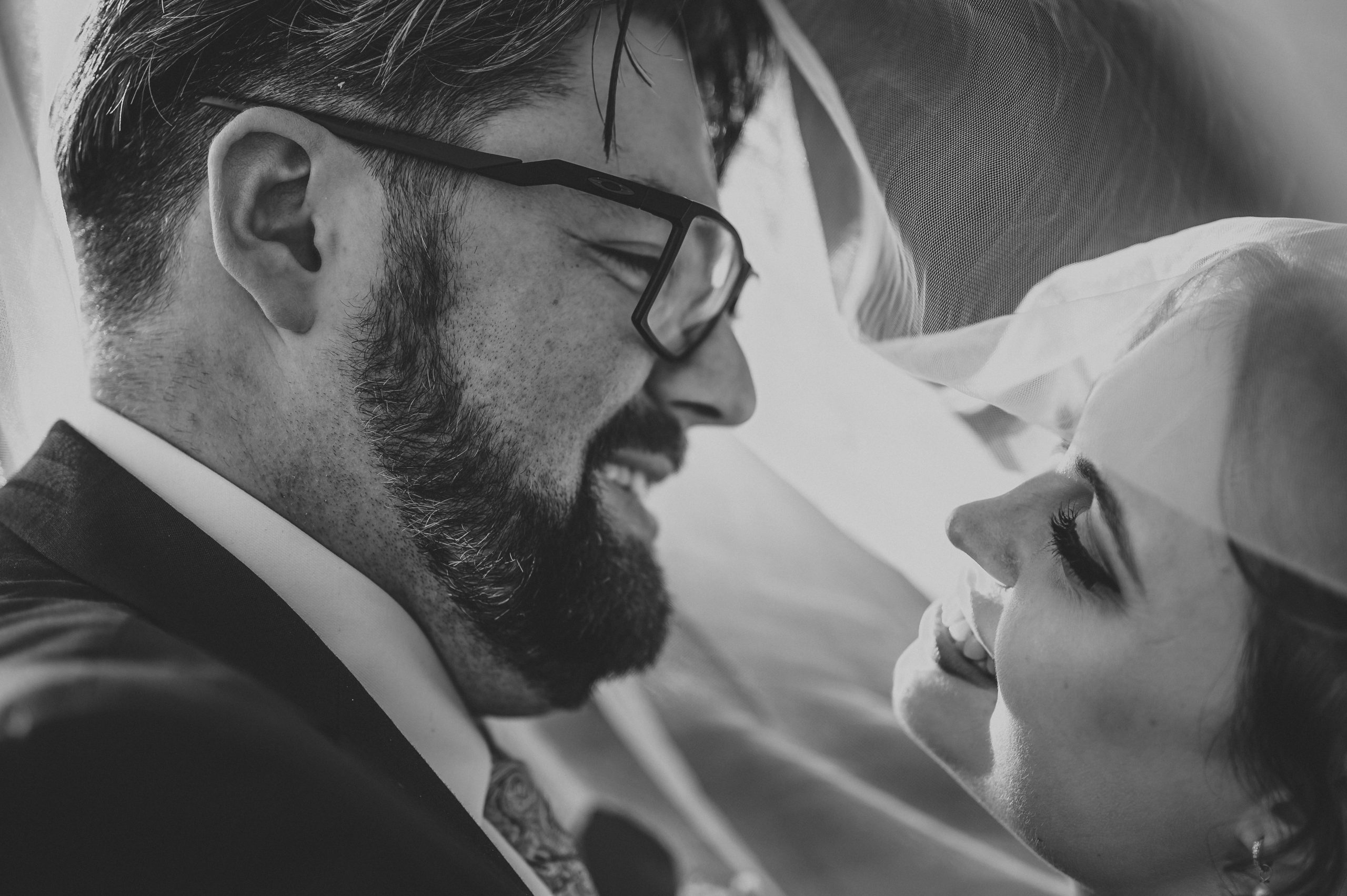 A black and white close-up of a bride and groom smiling at each other, captured by a Surrey wedding photographer. The groom, wearing glasses and a beard, gazes adoringly at the bride, whose face is partially covered by a veil, adding an enchanting touch to this romantic moment.